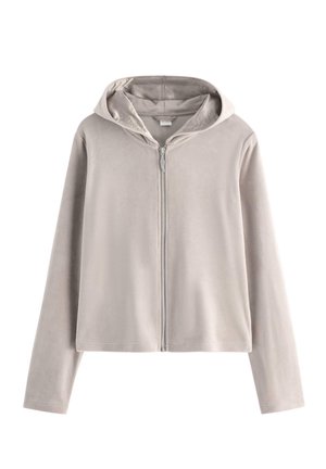 Tienermeisje staand, met een beige trainingspak, een roze hemdje met kant en zwarte ballerina's tegen een effen witte achtergrond.