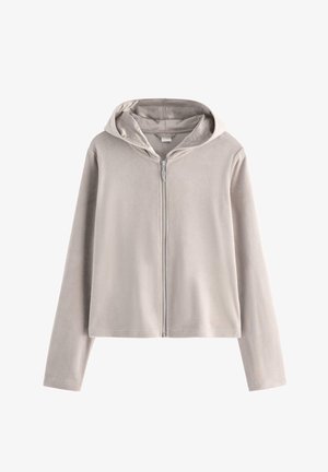 Teenagepige stående iført beige joggingdragt med lyserød top med blonderkant og sorte balletflade sko mod en ensfarvet hvid baggrund.