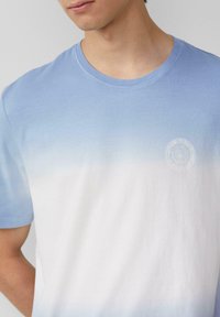Helles blauer Farbverlauf T-Shirt mit kurzen Ärmeln, das einen runden Ausschnitt hat und von hellblau zu Weiß übergeht, mit einem dezenten Logodetail.
