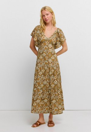 Vrouw met blond haar die een bruine patroon maxi-jurk met zwierige mouwen en bruine sandalen draagt, staand tegen een effen lichte achtergrond.