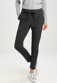 Calças cinzentas escuras com um corte descontraído, cinto atado, bolsos laterais e bainhas com punho, combinadas com sapatos prateados de deslizar. Textura suave, tecido liso.