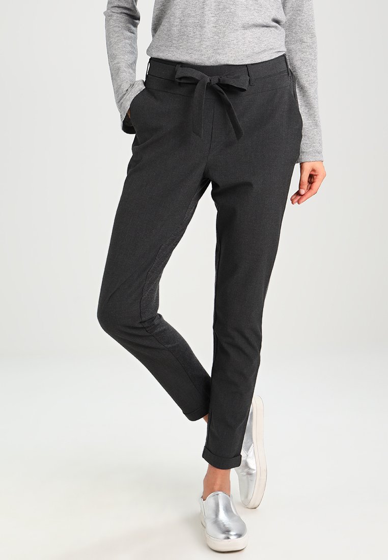 Calças cinzentas escuras com um corte descontraído, cinto atado, bolsos laterais e bainhas com punho, combinadas com sapatos prateados de deslizar. Textura suave, tecido liso.