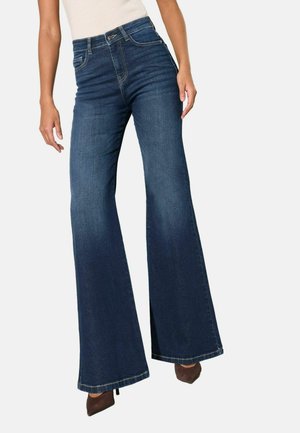 Naine, kes kannab kõrge vöökohaga tumelood siniseid lõhnavad teksaseid ja pruune teravate tõstukingadega, seisab ühevärvilise hele tausta ees.