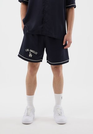 Uomo che indossa pantaloncini blu navy con la scritta "Los Angeles LA", sneakers bianche, calzini bianchi e una camicia abbottonata blu navy coordinata, su uno sfondo semplice.