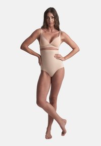 Donna in piedi a piedi nudi con le braccia sui fianchi, che indossa biancheria intima shapewear beige a vita alta e un reggiseno coordinato su uno sfondo neutro.