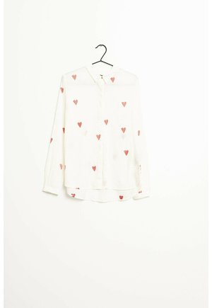 Biała bluzka z długim rękawem z czerwonymi wzorami serc, zapinana z przodu na guziki, z kołnierzykiem, wisząca na czarnym wieszaku na białym tle.