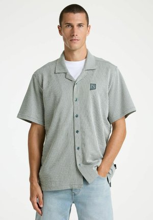 Man draagt een grijs-wit verticale gestreept overhemd met korte mouwen over een wit T-shirt, en lichtblauwe spijkerbroek, staand tegen een effen achtergrond.