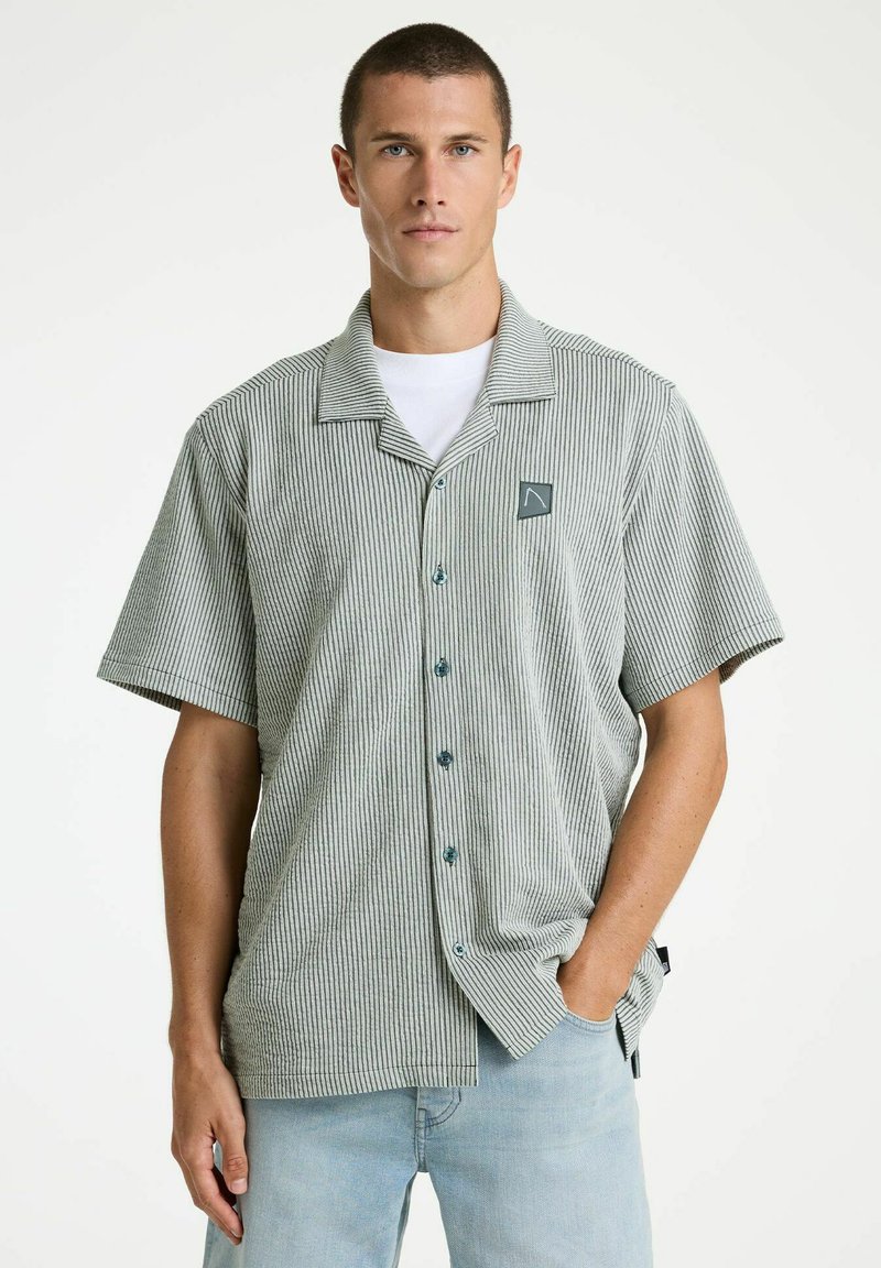 Hombre con camisa de botones de manga corta a rayas verticales grises y blancas sobre camiseta blanca, y vaqueros azul claro, de pie contra un fondo liso.