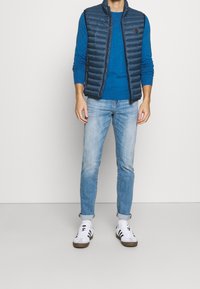 Doudoune sans manches bleu marine portée sur un pull bleu, associée à un jean bleu clair et des baskets blanches avec des accents noirs. Tissu lisse et coupe ajustée.