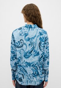 Camicia blu con motivo paisley, maniche lunghe, colletto e schiena scoperta. Presenta disegni intricati in diverse sfumature di blu e bianco.
