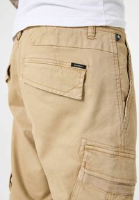 Beige cargobroek gemaakt van katoen, met meerdere zakken, stiksels en een label op de achterzak. Gelaagd stof.
