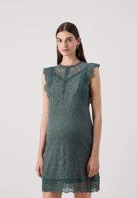 Zelena čipkasta haljina s ušivenim rukavima u obliku valova i cvjetnim uzorkom, koja ima uski kroj i teksturirani rub. Elegantna i detaljno osmišljena.