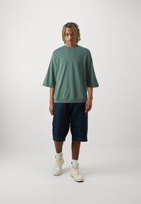 Turkise oversized sweatshirt med korte ærmer, parret med mørke denim shorts til knæene. Hvide træningssko fuldender looket.