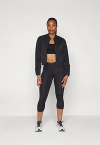 Črna kratka jakna v kombinaciji s črnim športnim modrčkom in črnimi kapri legicami, z nežnim logotipom na nogi in teksturirano tkanino.