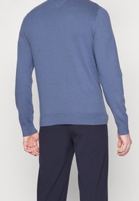 Sweter z dekoltem w serek w jasnoniebieskim dzianinowym materiale z prążkowanymi mankietami i dołem, w zestawieniu z ciemnymi spodniami. Widok z tyłu pokazuje teksturowany wzór.