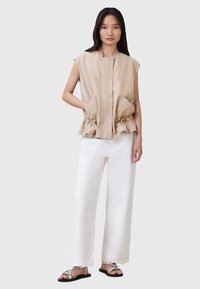 Gilet beige senza maniche con fronte plissettato e vita elastica, dotato di due tasche frontali. Abbinato a pantaloni bianchi a gamba larga e sandali.