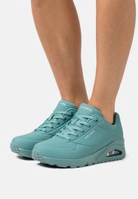 Teal Skechers sneakers with a smooth synthetic upper, lace-up design, perforated detailing, and a visible air cushion in the sole.