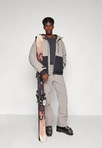 Síruha szürke és fekete dzsekivel, szürke nadrággal és síálarccal. Egy bézs és sötétrózsaszín mintájú sílécet tart, fekete csizmával.