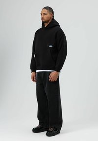 Zwarte hoodie met een voorzak en een wit logodetail. Gecombineerd met losvallende zwarte spijkerbroek en zwarte veter schoenen. Minimalistisch ontwerp.