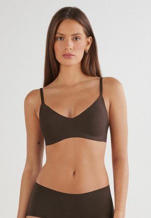 Mujer con cabello largo y castaño lleva un sujetador sin costuras marrón oscuro y ropa interior a juego frente a un fondo blanco liso.