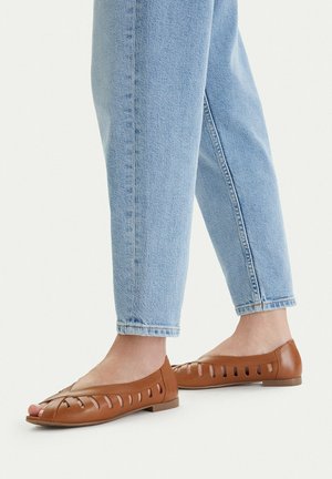 Osoba ubrana w jasnoniebieskie dżinsy i brązowe skórzane płaskie sandały z wycięciami, stojąca na jednolitym jasnym tle.