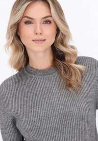 Grå ribbet sweater med høj krave. Blød tekstur, tætsiddende design. Model med bølget hår poserer foran en ensfarvet hvid baggrund.