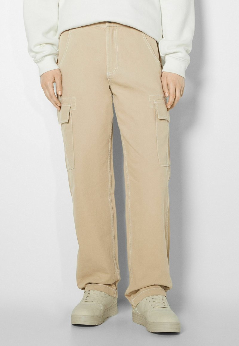 Pantalon cargo beige avec poches latérales, coupe droite, coutures contrastantes et tissu en coton résistant. Porté avec des baskets de couleur claire.