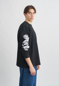 Μαύρο κοντομάνικο μπλούζα με ριμπ ύφανση που διαθέτει λευκό γραφικό σχέδιο στο αριστερό μανίκι, σε συνδυασμό με μπλε τζιν παντελόνι.