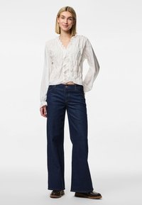 Blusa branca com golas franzidas de manga longa, combinada com jeans de perna larga de cintura alta em azul escuro. Tecido texturizado e corte relaxado, com sapatos castanhos.