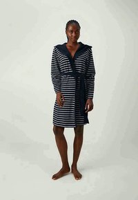 Kobieta stoi boso, ubrana w granatowo-białą paskowaną kąpielówkę z kapturem związaną w talii, na jednolitym jasnym tle.