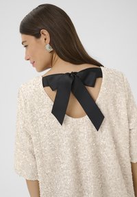 Mirdzošs beidzis topiņš ar apaļu kakla izgriezumu, atvērtu aizmuguri un melnu satīna loku detaļu. Īsas piedurknes un brīvs piegriezums.