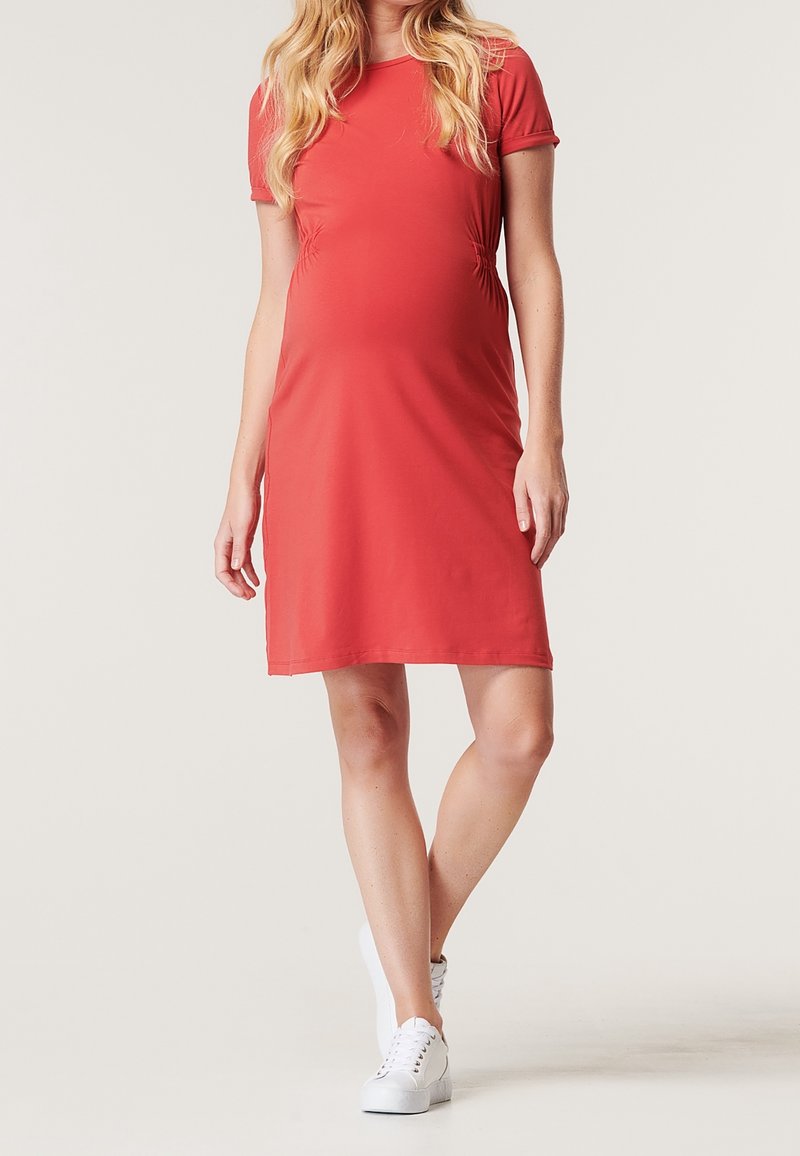 Femme enceinte portant une robe de maternité corail à manches courtes avec des côtés froncés et des baskets blanches, debout devant un fond uni.