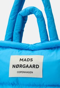 Blå vadderad väska med två handtag och en vit etikett som visar "MADS NØRGAARD COPENHAGEN" i svarta bokstäver.