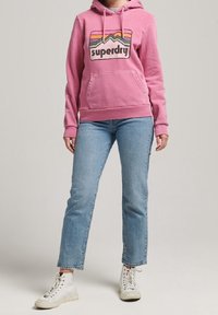 Felpa con cappuccio rosa e logo grafico, abbinata a jeans dritti azzurri chiari e sneakers bianche. Design semplice e casual.