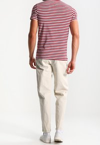 Hombre de pie de espaldas, vistiendo una camisa de manga corta a rayas rojas y blancas, pantalones beiges, calcetines blancos y zapatillas blancas.