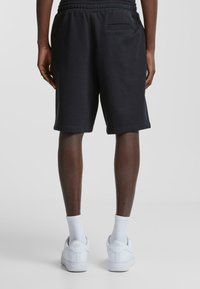 Pantalones cortos de algodón negros con una cinturilla elástica, bolsillos laterales y un único bolsillo trasero. Combinados con zapatillas blancas y calcetines.