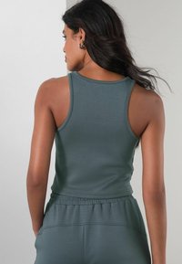 Tumši zaļš, piegriezts bezpiedurkņu krekls ar apaļu kakla izgriezumu un platām plecu lencēm. Gluda tekstūra, minimālas šuves, kopā ar atbilstošiem jogger biksēm.