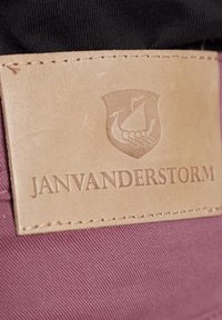 Beige leren patch met geperst schild en scheepsontwerp, genaaid op donkerpaarse stof. Tekst luidt "JANVANDERSTORM."