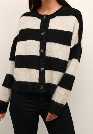 Femme portant un cardigan boutonné à larges rayures horizontales noires et blanches à manches longues et un pantalon noir.