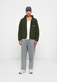 Grønn fleecejakke med svart lomme, grå cargo-bukser og hvite sneakers. Modellen står mot en ensfarget hvit bakgrunn.