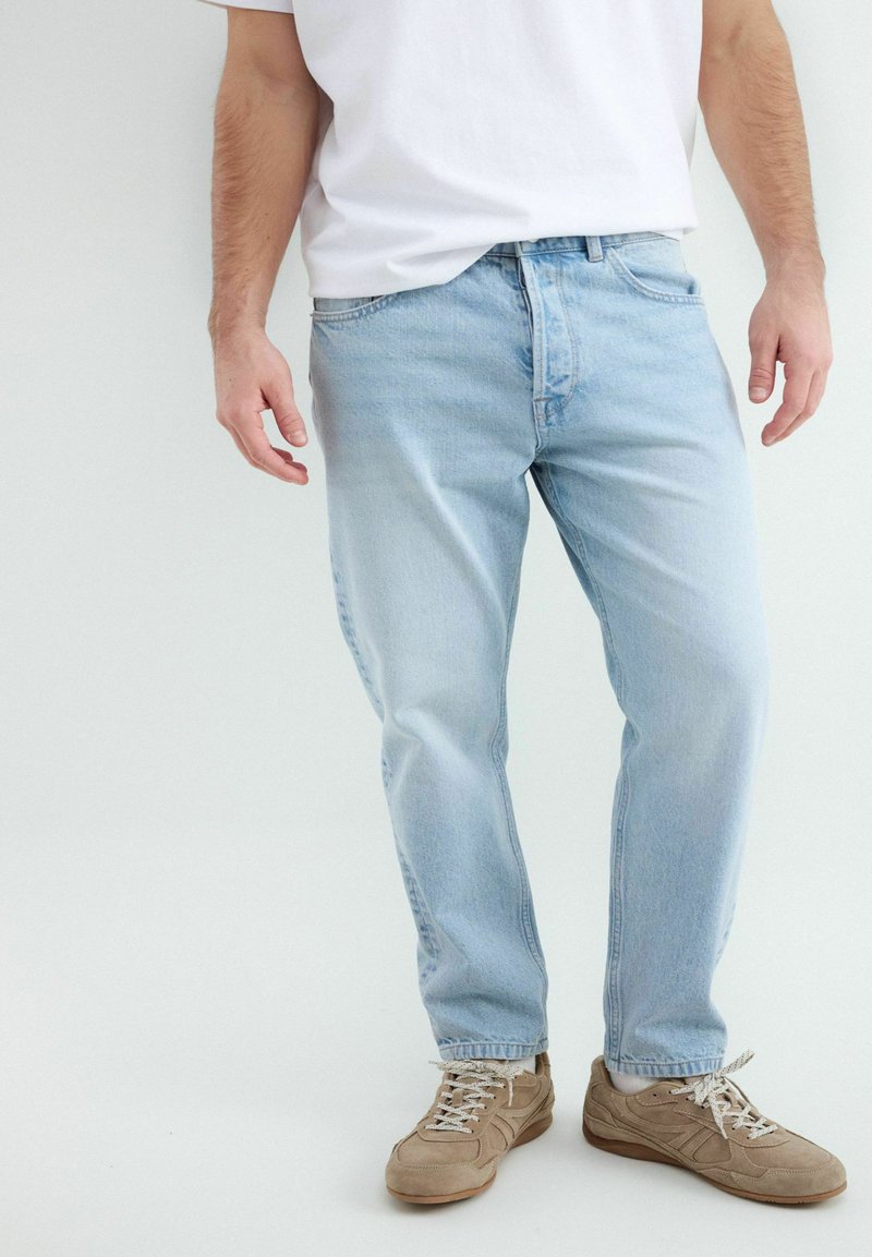 Homme portant un jean bleu clair, des chaussettes blanches, des baskets beige et un t-shirt blanc sur un fond blanc uni.