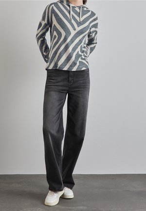 Osoba v mikině s šedobílým geometrickým vzorem, černých rovně střižených džínách a bílých teniskách stojí před jednobarevnou zdí.