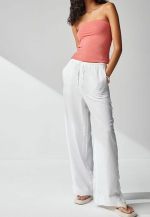 Jonge vrouw met lang haar die een koraalkleurige strapless top, witte wijde broek met trekkoord en beige sandalen draagt, staat met handen in de zakken.