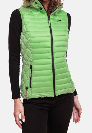 Femme portant un gilet sans manches matelassé vert vif avec une fermeture éclair à l'avant, par-dessus un chemisier noir à manches longues et un pantalon noir.