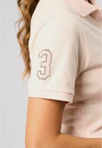 Lichtroze poloshirt met een kraag, voorzien van een opvallend geborduurd nummer "3" in een donkere schaduw op de linkermouw.