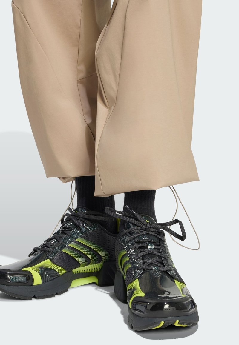 Chaussures de sport noires et vert néon avec une tige en mesh, des accents texturés et des lacets noirs. Portées avec un pantalon beige ample et des chaussettes noires.