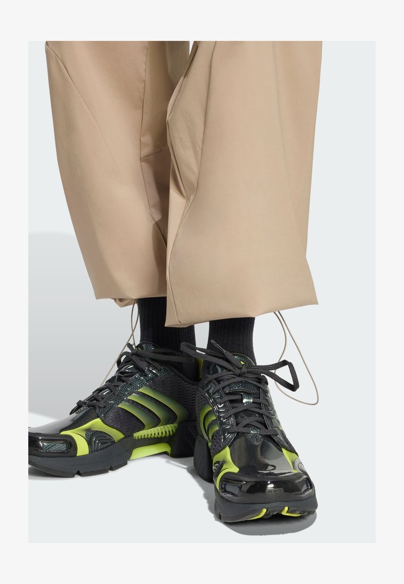 Zapatillas deportivas negras y verde neón con parte superior de malla, detalles texturizados y cordones negros. Usadas con pantalones beige holgados y calcetines negros.