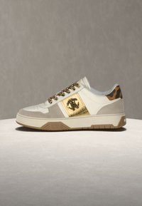 Baskets basses avec des panneaux blancs, beige et dorés, des lacets à imprimé léopard et un motif léopard au talon sur un fond uni.