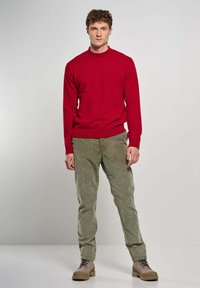 Giovane uomo in piedi, rivolto in avanti, indossa un maglione rosso, pantaloni in velluto a coste verde e stivali beige con lacci, su uno sfondo chiaro e semplice.