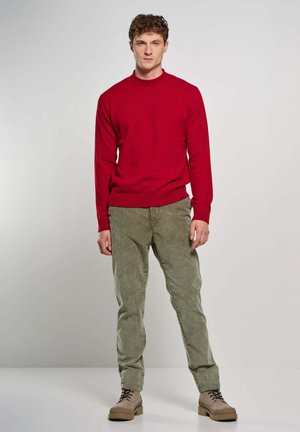 Young man stands facing forward wearing a red sweater, green corduroy pants, and beige lace-up boots against a plain light background.